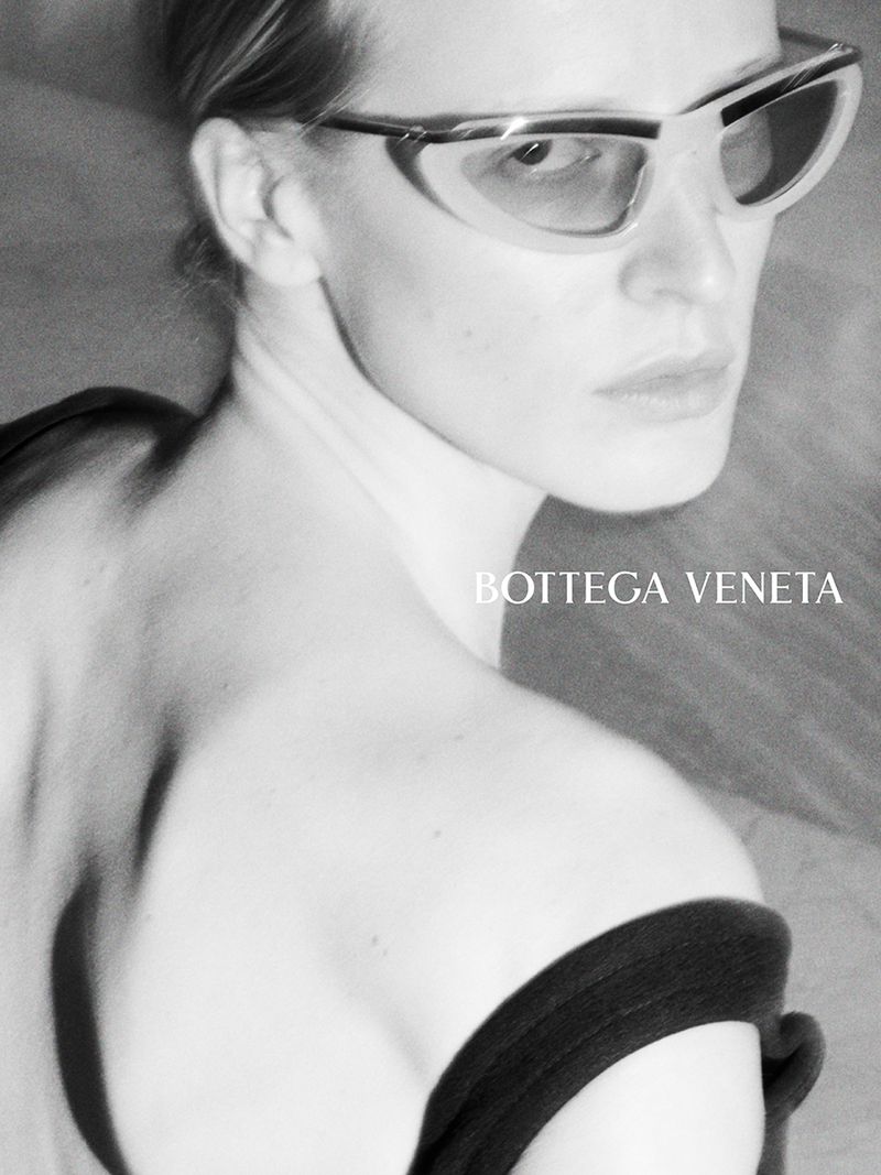 Black and white close-up of a model looking over her shoulder wearing semi-transparent cat-eye sunglasses and an off-shoulder dark top.
