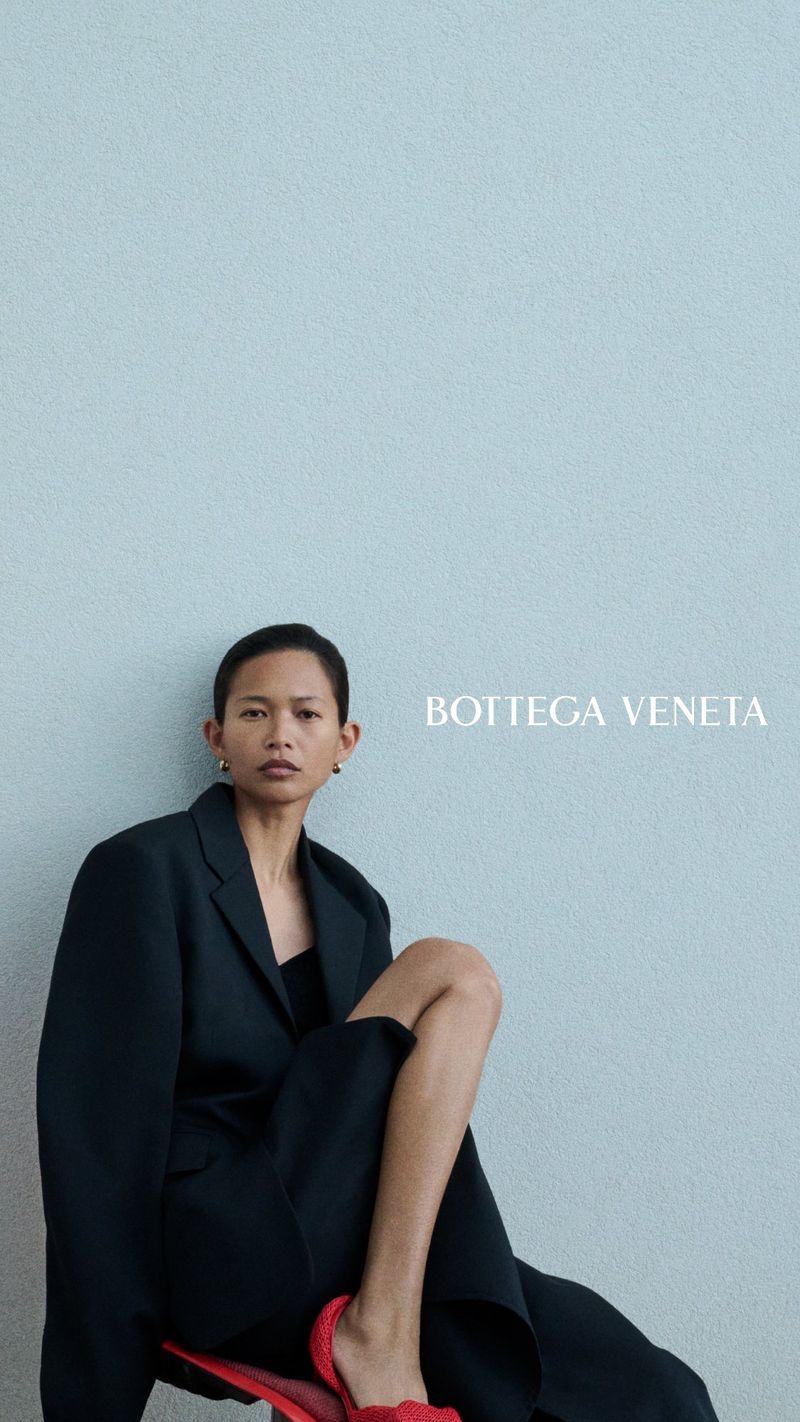 Model in an oversized black blazer and red shoes seated against a pale blue textured wall with Bottega Veneta text.