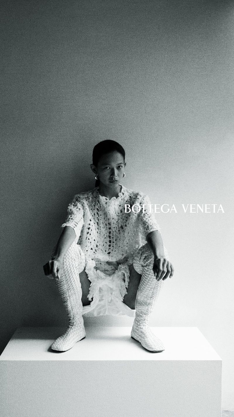 Model Karmay Ngai crouching on a plinth in an openwork white knit dress and tall boots for the Bottega Veneta campaign.