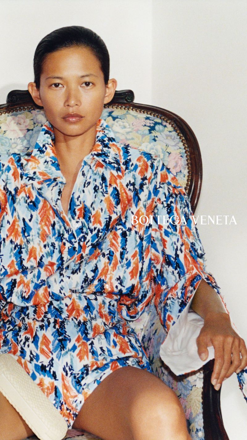 Karmay Ngai wearing a blue and orange patterned shirt seated on a floral vintage armchair against a white background.