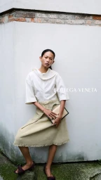 Model Karmay Ngai poses in a white blouse, beige fringed skirt, and Bottega Veneta handbag against a white wall corner.
