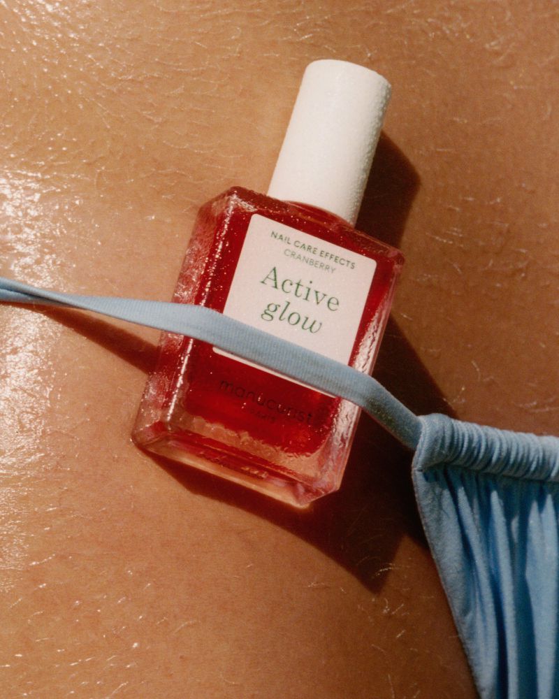 Close-up of red nail polish bottle on tanned skin beside a light-blue swimsuit. 'Active Glow' text on the label.