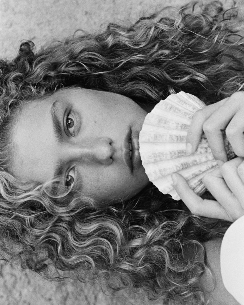 Monochrome portrait of a young woman with curly hair holding a large seashell against a neutral sandy background, focusing on natural beauty.