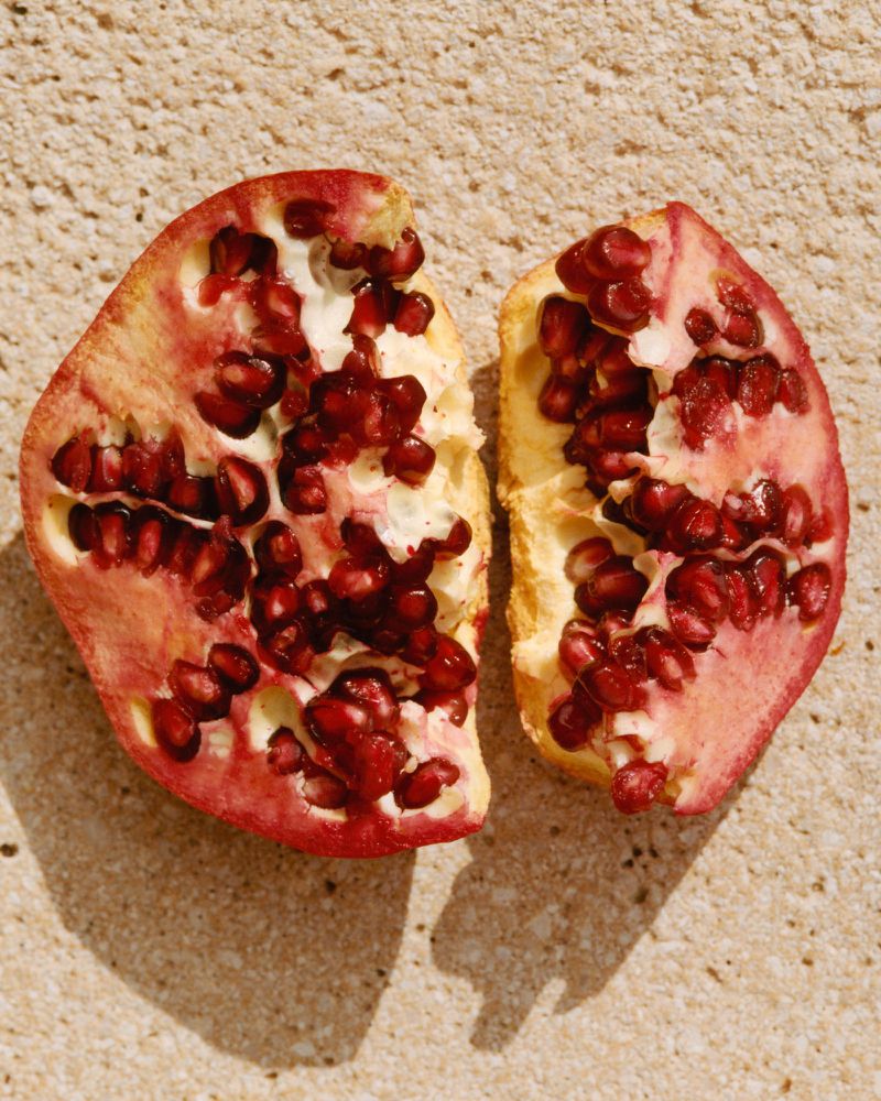 Overhead shot of a halved pomegranate on a textured tan surface, seeds visible, illuminated by bright, diffused natural light.
