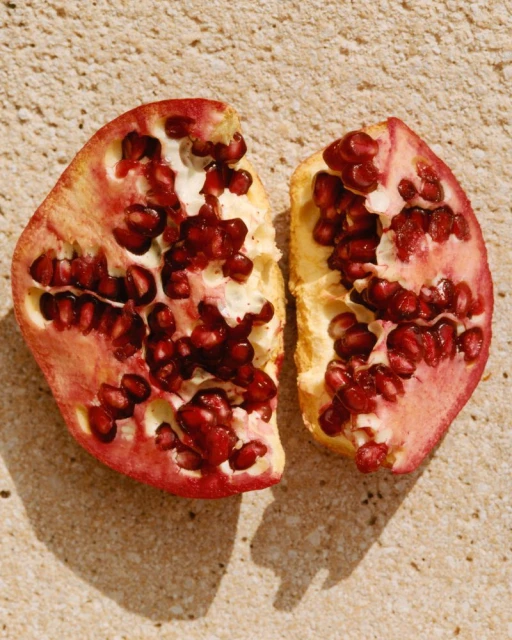 Overhead shot of a halved pomegranate on a textured tan surface, seeds visible, illuminated by bright, diffused natural light.
