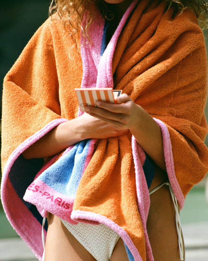 Woman wrapped in an orange, pink, and blue towel holds a small striped compact case in an outdoor setting with natural light.