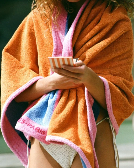 Woman wrapped in an orange, pink, and blue towel holds a small striped compact case in an outdoor setting with natural light.
