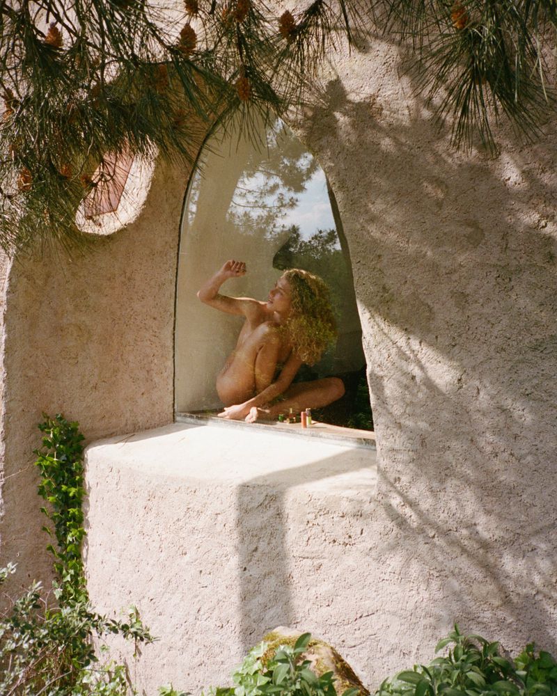 Nude fair-skinned woman with curly hair sits inside a stone archway window, partially framed by tree branches and foliage, outdoor scene.