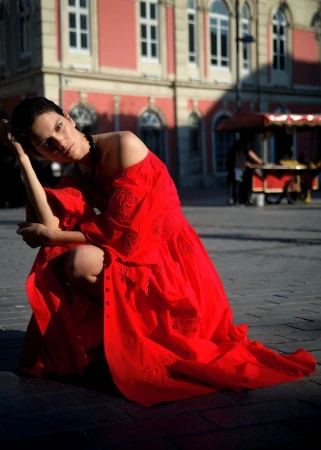 Adult woman in a bright red dress is kneeling outdoors with buildings and a food cart visible in the background, during daylight hours.