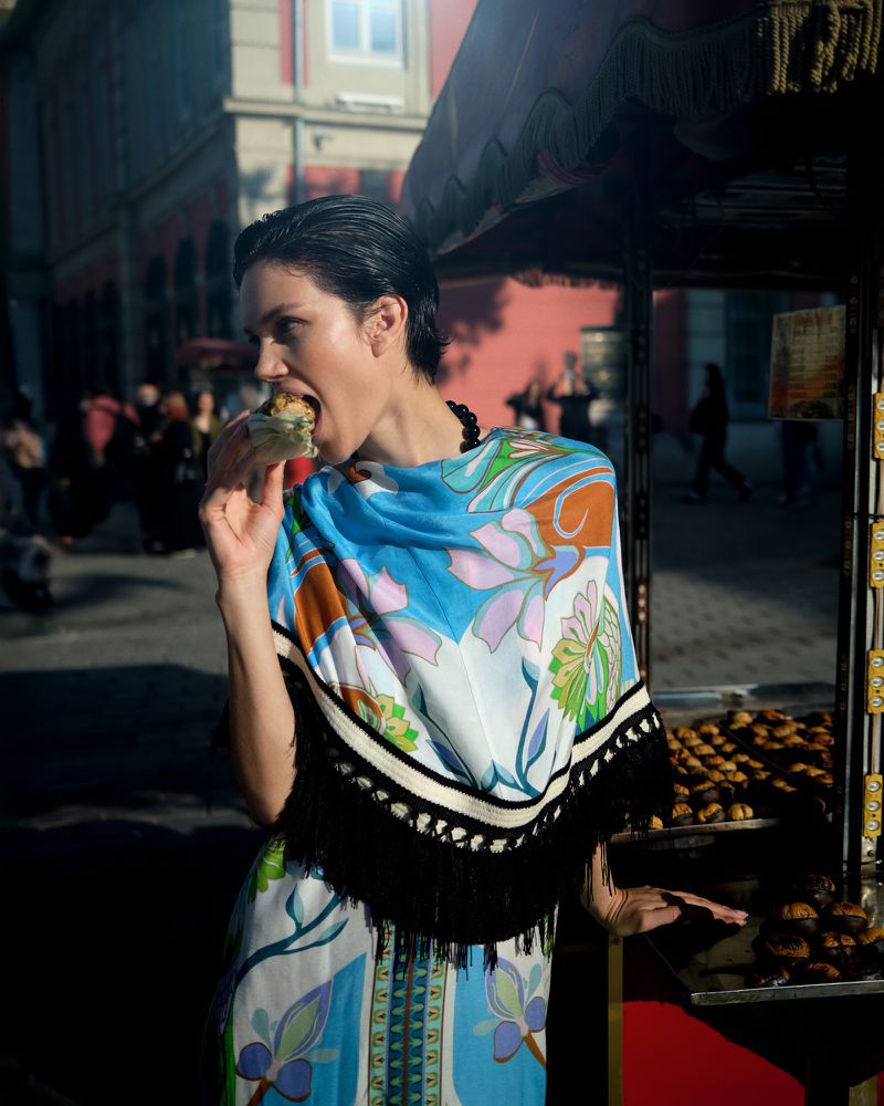 Young woman in a patterned dress eating food at a street vendor in a city setting, showcasing urban life and culinary culture.