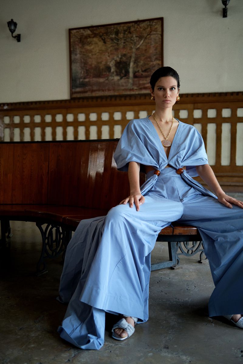 A woman sits on a wooden bench in a light blue outfit and jewelry, posing in front of a wall with a painting and sconces.