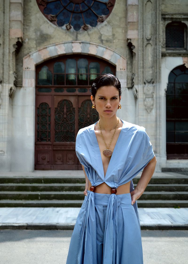 A woman stands in front of a stone building wearing an open light blue outfit, a gold necklace, and large gold earrings. 
