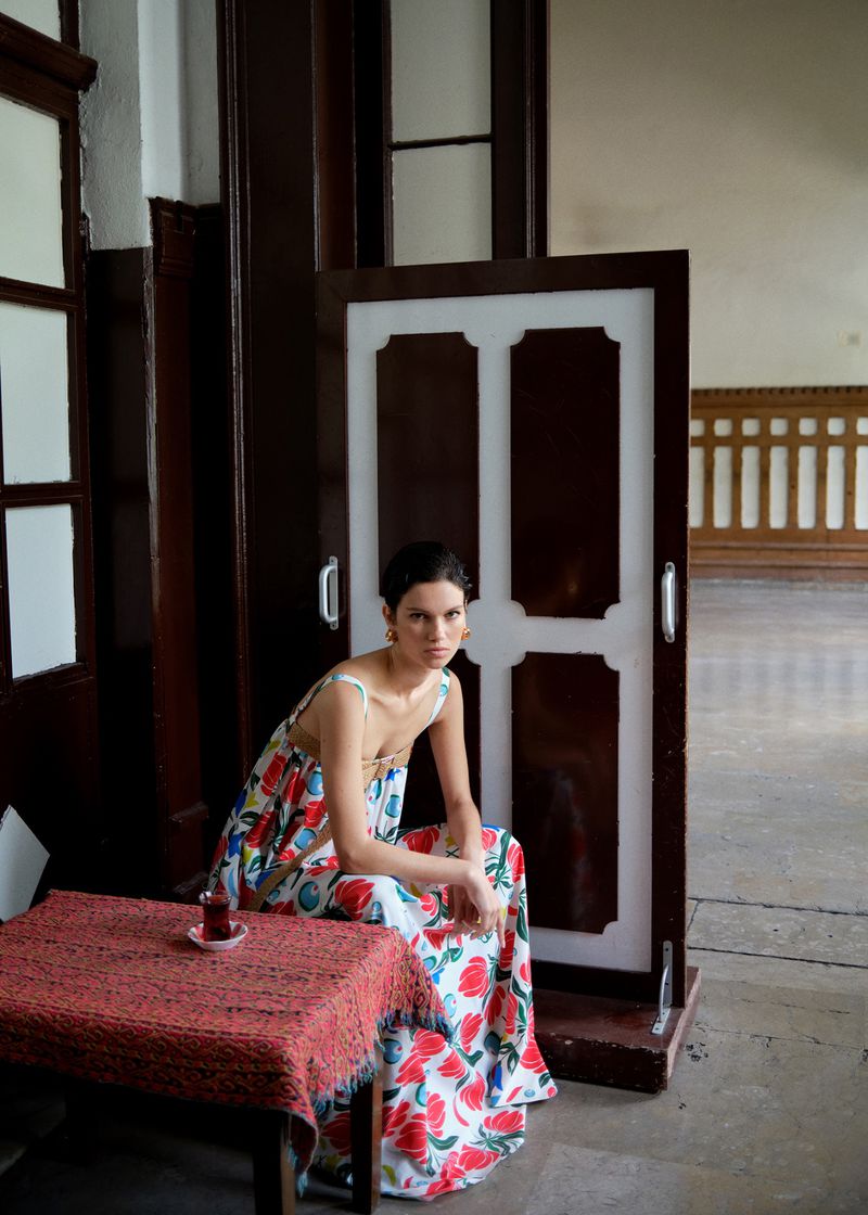 Adult woman wearing floral dress is seated at a red table in an indoor setting near a white and brown door, with natural light.