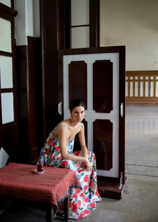 Adult woman wearing floral dress is seated at a red table in an indoor setting near a white and brown door, with natural light.