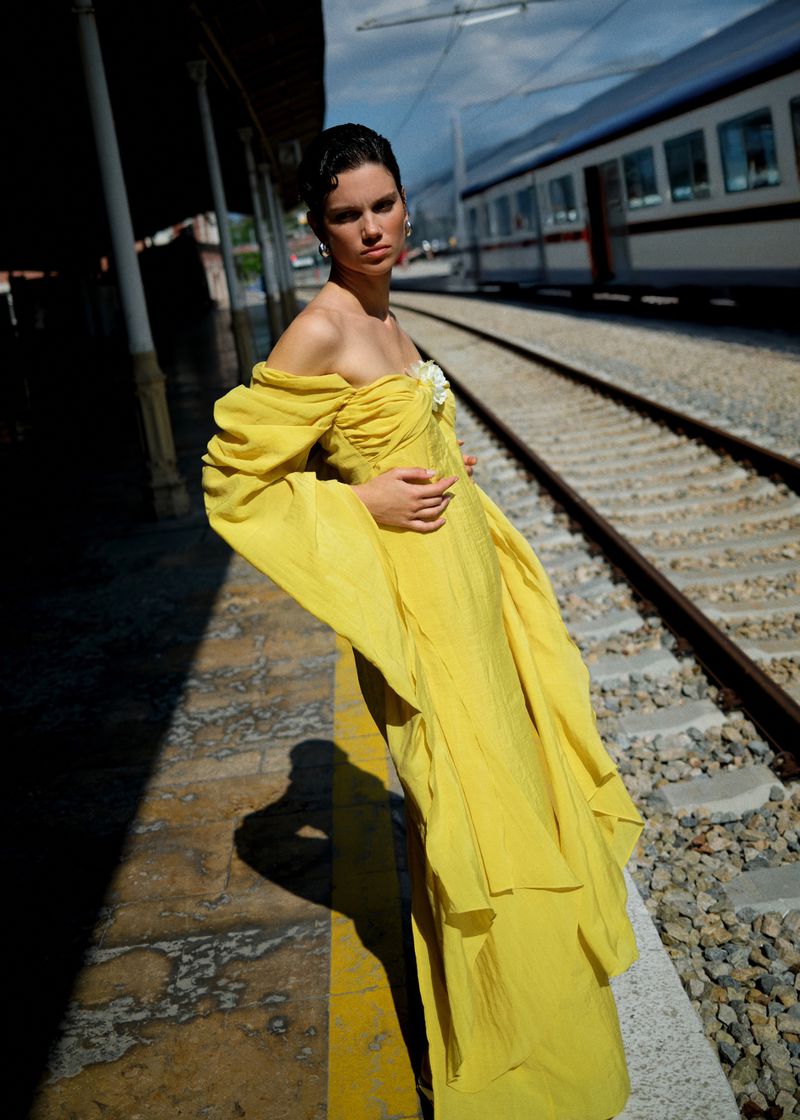 A stylish woman in a flowing yellow gown stands on a train platform with a passenger train in the background under a partly cloudy sky.