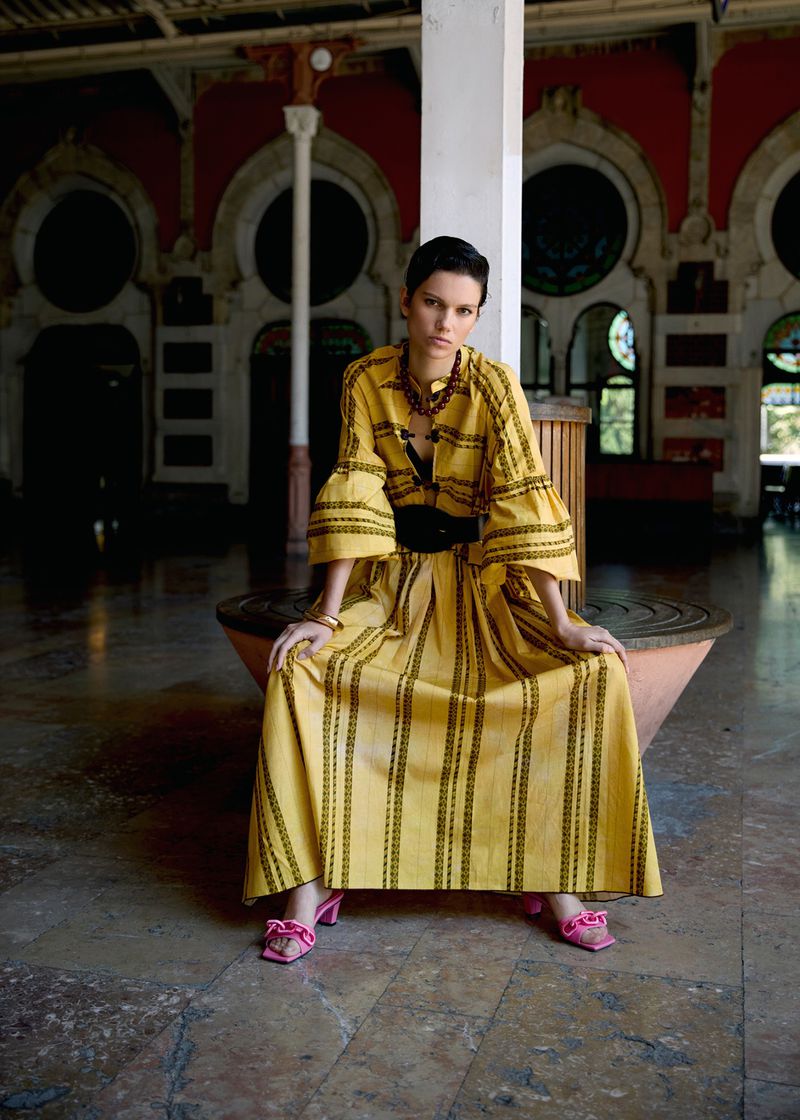 Young woman in a yellow dress and pink heeled shoes poses sitting down in an indoor location with arched window details in the background.