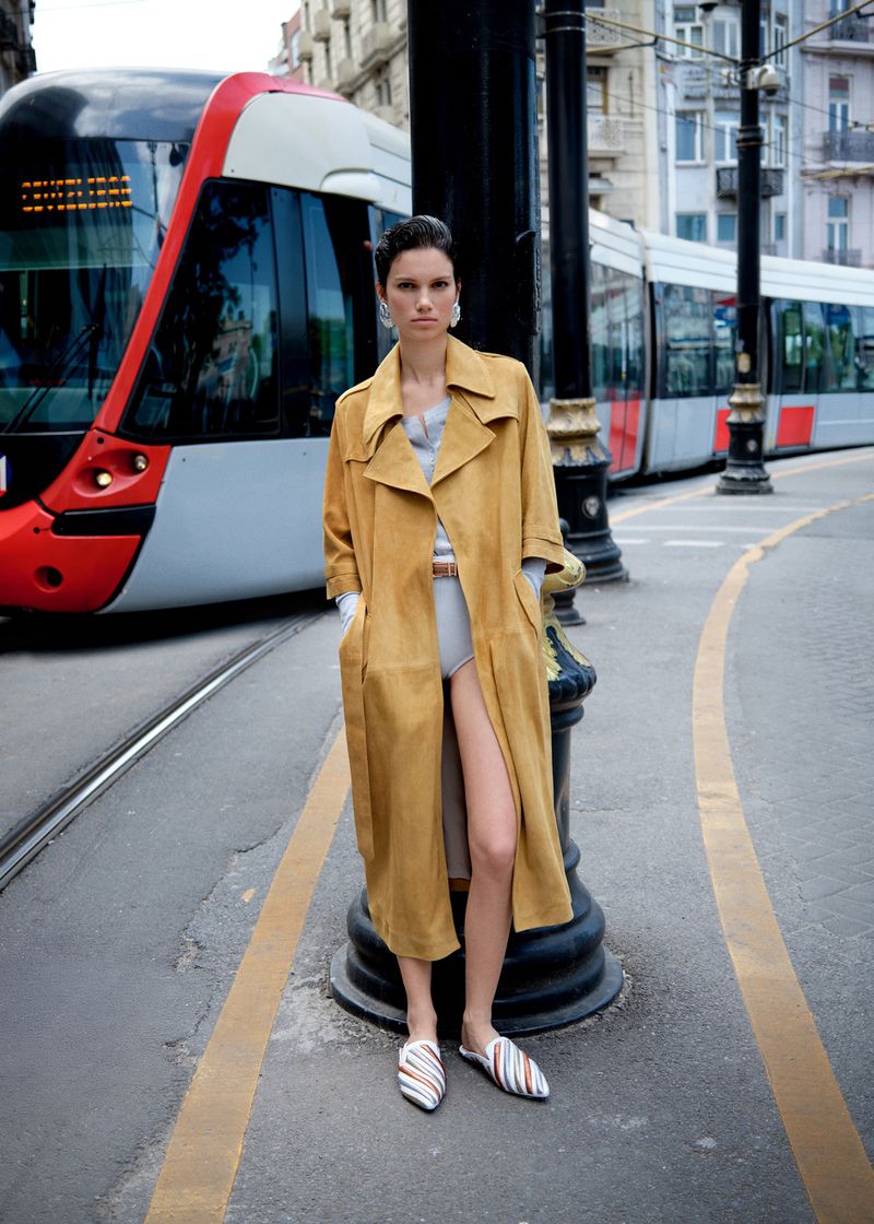 Fashion editorial featuring a woman in a long coat posing on a city street with a tram in the background, creating a stylish urban scene.