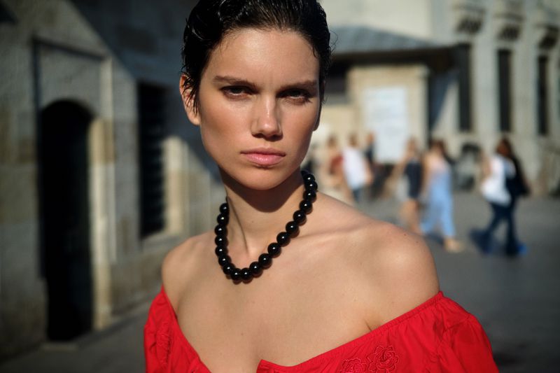 Portrait of a woman with dark hair and a red off-the-shoulder top, wearing a black bead necklace, outdoors in an urban environment.