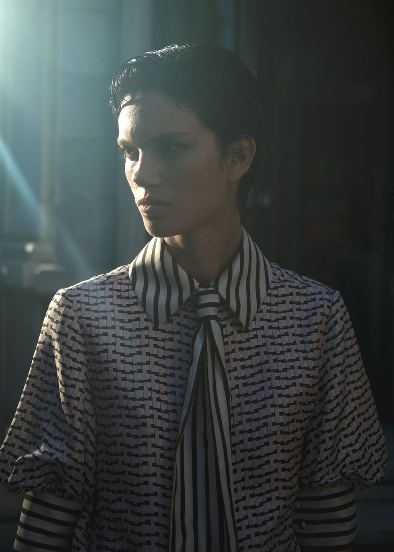 Portrait of a female model in a patterned blouse with a striped scarf, partially lit with sun flares in a shadowed setting.