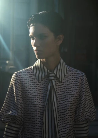 Portrait of a female model in a patterned blouse with a striped scarf, partially lit with sun flares in a shadowed setting.