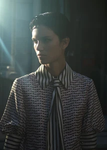 Model with short dark hair wearing a patterned top with a striped collar and tie, lit by dramatic beams of light.