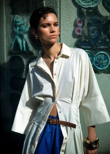 Model in white buttoned shirt and blue skirt with a brown belt outdoors against a textured tiled wall for Vogue Turkey.