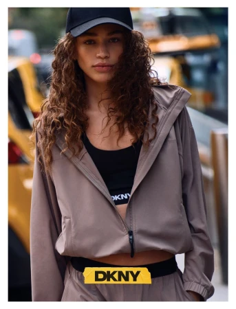 Model wearing a taupe zip-up windbreaker jacket, black sports bra, and black cap on a city street.