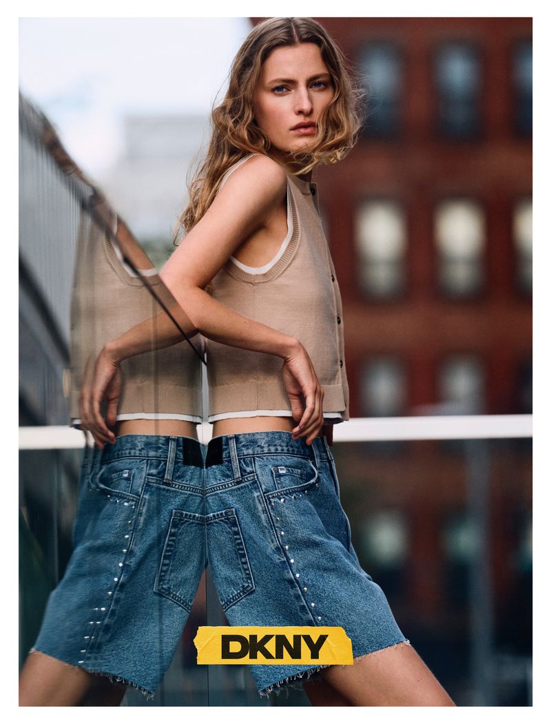 Model Felice Nova Noordhoff wearing a beige sleeveless top and denim shorts leans against a reflective glass railing for DKNY.
