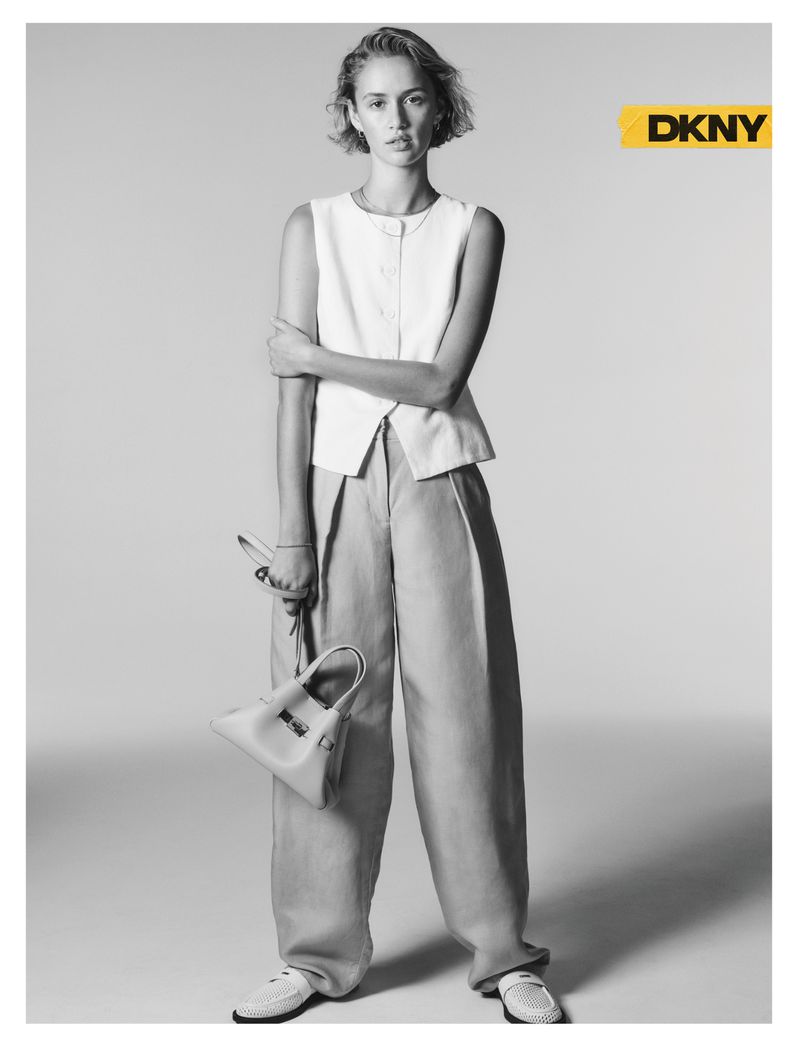Quinn Mora in a white sleeveless vest and pleated wide-leg trousers holding a structured handbag for the DKNY Spring 2026 campaign.