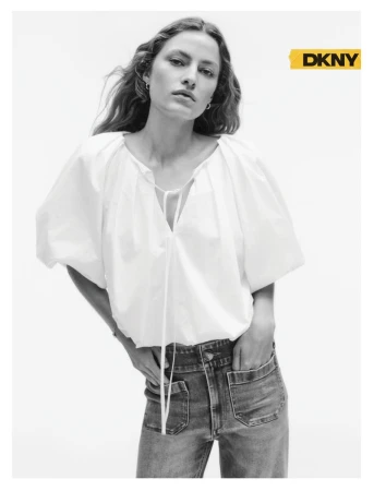 Model wearing a white puff-sleeve tie-front blouse and denim jeans with hands in pockets against a plain white studio background.