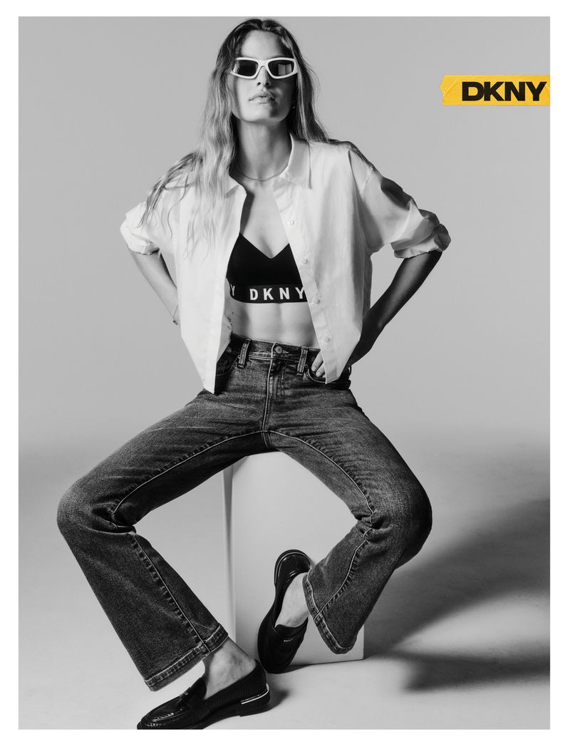 Model wearing white sunglasses, an open white shirt, DKNY branded bra, and denim jeans seated against a studio backdrop.