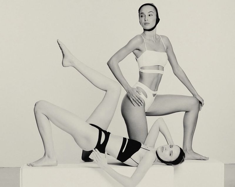 Two female models posing in swimwear against a plain studio background, with one standing and one lying on blocks.