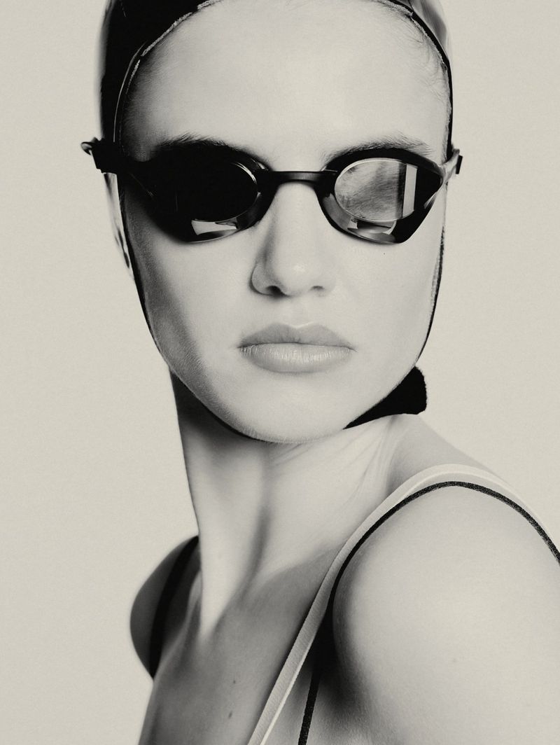Monochrome portrait of a female swimmer with goggles and a swim cap, captured in a studio setting with soft lighting. The focus is on her face.