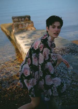 Woman with short dark hair wearing a black floral print dress crouching near a stone pier by the water.