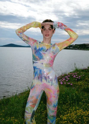 Model wearing a multi-color patterned full-body jumpsuit posing near a lake with grassy shoreline and overcast sky