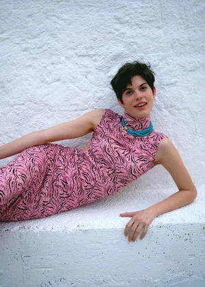 Model with short dark hair in a pink patterned dress lying against a textured white wall for 5ELEVEN Magazine
