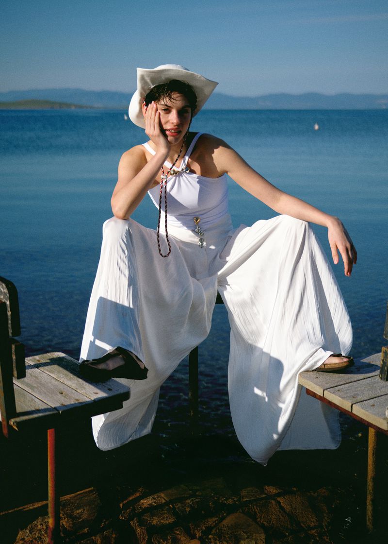 Woman in white outfit and hat sitting at lakeside with blue water, mountains, and clear sky in the background on a sunny day.