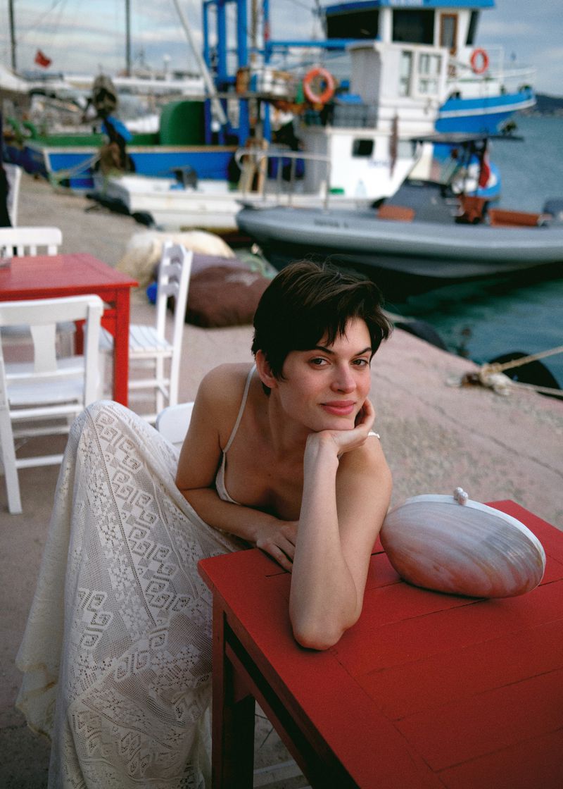 Brunette woman in white dress seated at red table with white purse, set outdoors near harbor with boats in the background on sunny day.