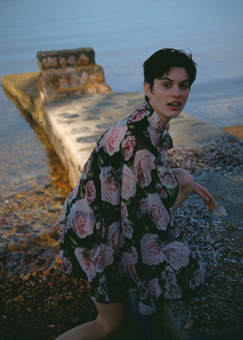 A young woman with short dark hair poses outdoors in a floral dress, kneeling on a stone surface near water with a concrete structure behind.
