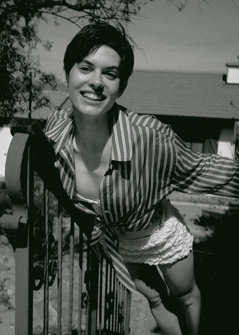 Black and white portrait of a smiling adult female in a striped shirt leaning on a fence outdoors on a sunny day, vintage aesthetic.