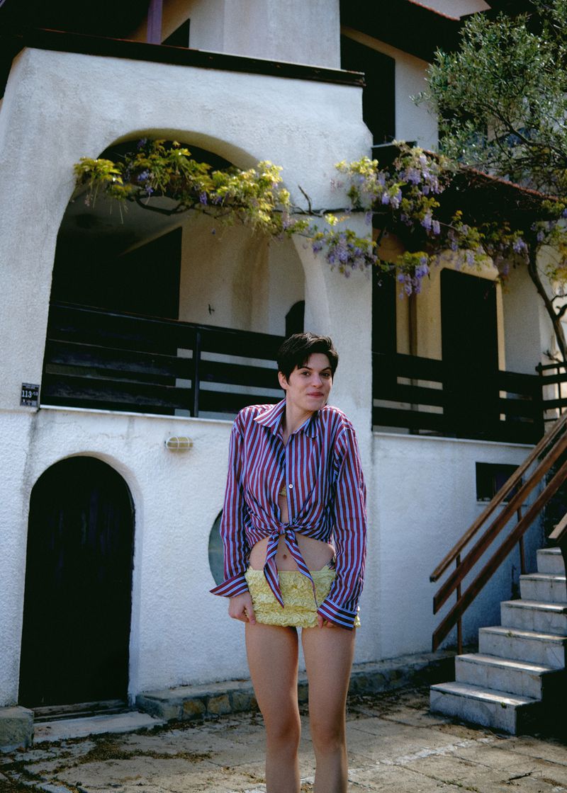 Young adult woman in a striped button-down shirt stands outside a stucco house with wisteria and stairs in a sunny, eye-level full shot.
