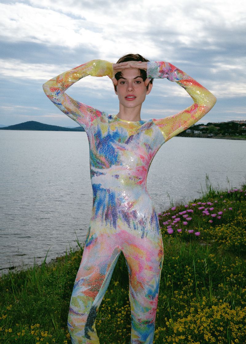 Adult female stands in a full-body multicolored patterned spandex suit with hands above her head in a green, grassy, lakeside setting.