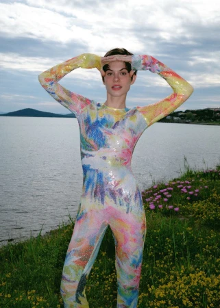 Adult female stands in a full-body multicolored patterned spandex suit with hands above her head in a green, grassy, lakeside setting.