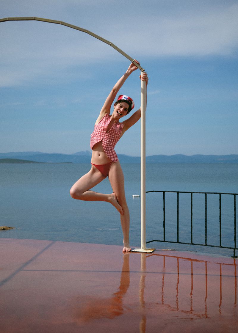 Woman in red-and-white checkered vest and red bikini bottom poses on an outdoor platform against a sea and sky backdrop.