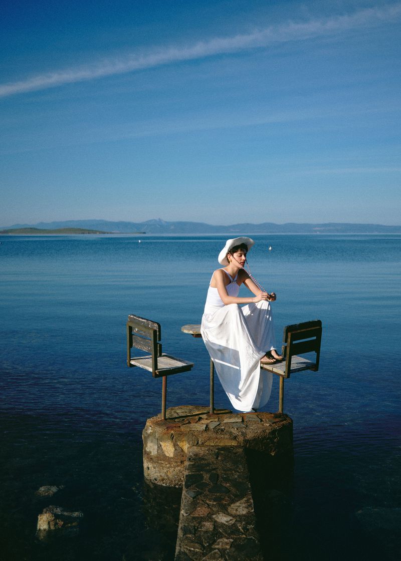 Woman in white dress and sunhat sits at table on stone pier overlooking a blue lake with distant mountains under a clear sky.