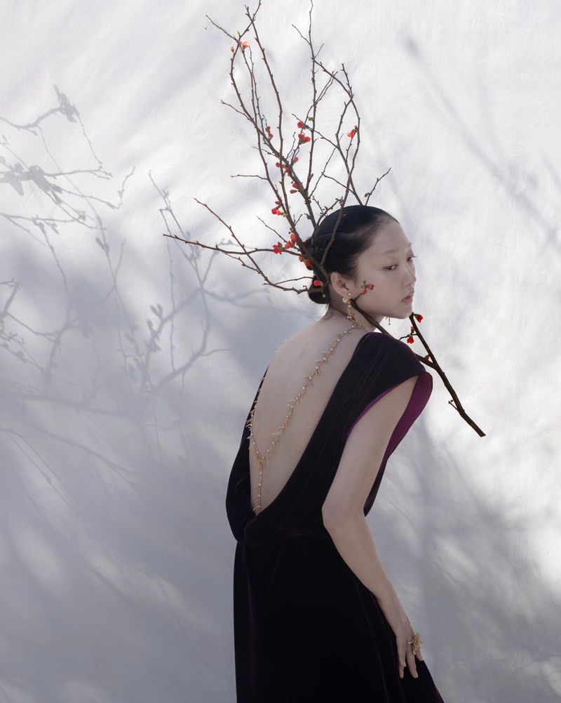 Asian woman in low-back formal gown, styled with branches and berries, poses in front of a wall casting tree branch shadows.