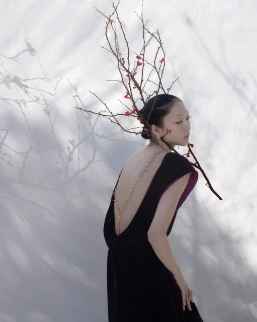 Asian woman in low-back formal gown, styled with branches and berries, poses in front of a wall casting tree branch shadows.