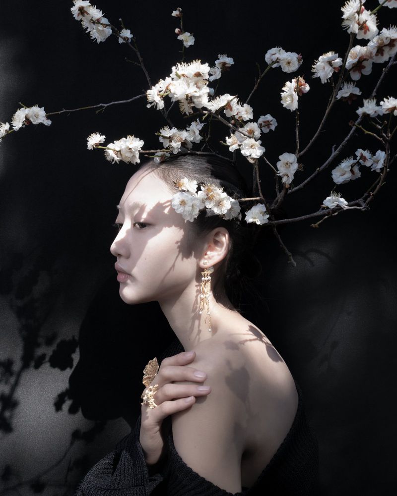 Portrait of a serene adult female with white blossoms in her hair, adorned with jewelry, set against a contrasting black background.