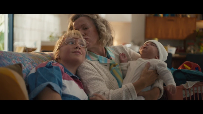 A heartwarming scene captures an elderly woman kissing a blond child on the head, while she's holding a baby with a white hat indoors.