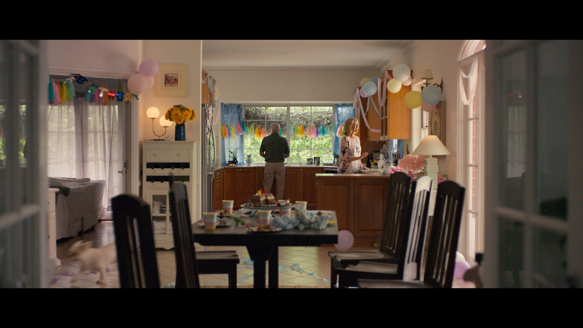 Interior shot of a home after a party, featuring two adults in the kitchen, party decorations, balloons, and a cluttered table.